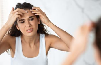 Young upset woman examining skin on her forehead in fron of a mirror.